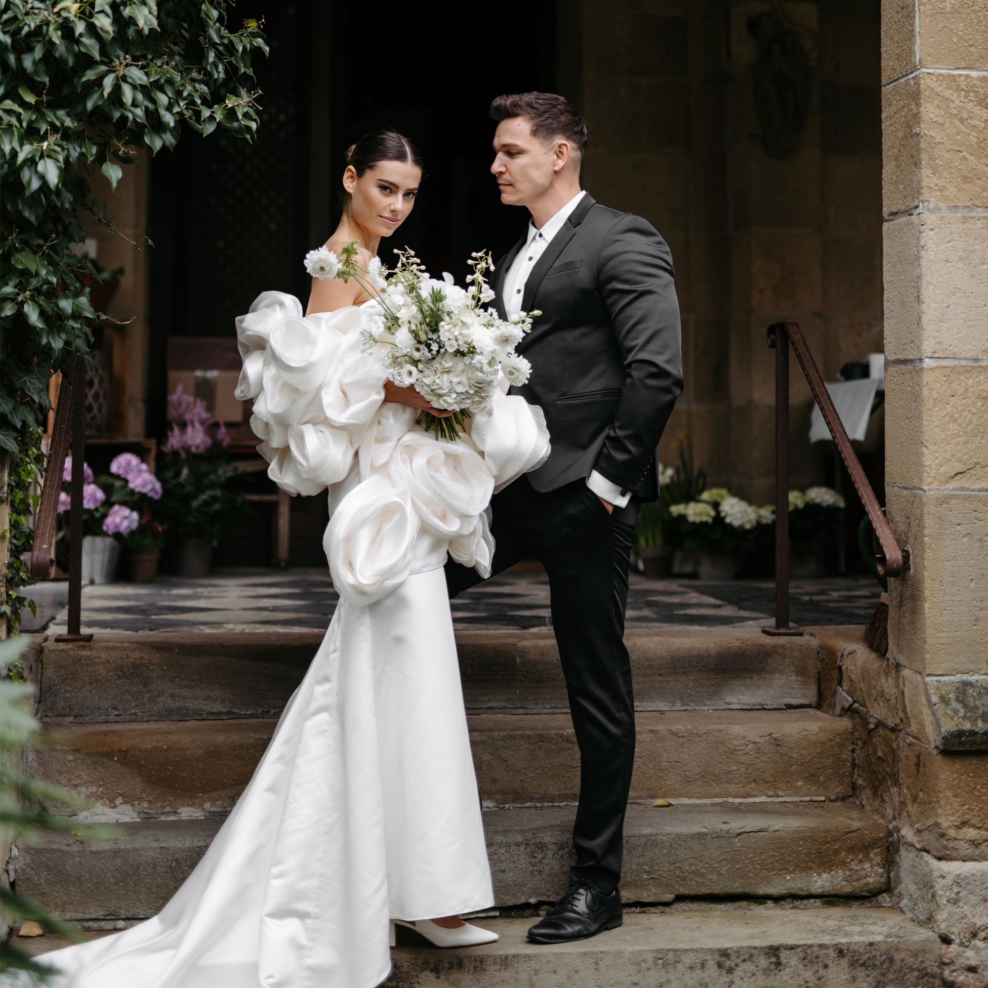 Braut in auffälligem, weißen Kleid und Brautstrauß aus weißen Blumen steht mit ihrem Bräutigam im dunklen Anzug auf einer Steintreppe vor einem alten Gebäude.
