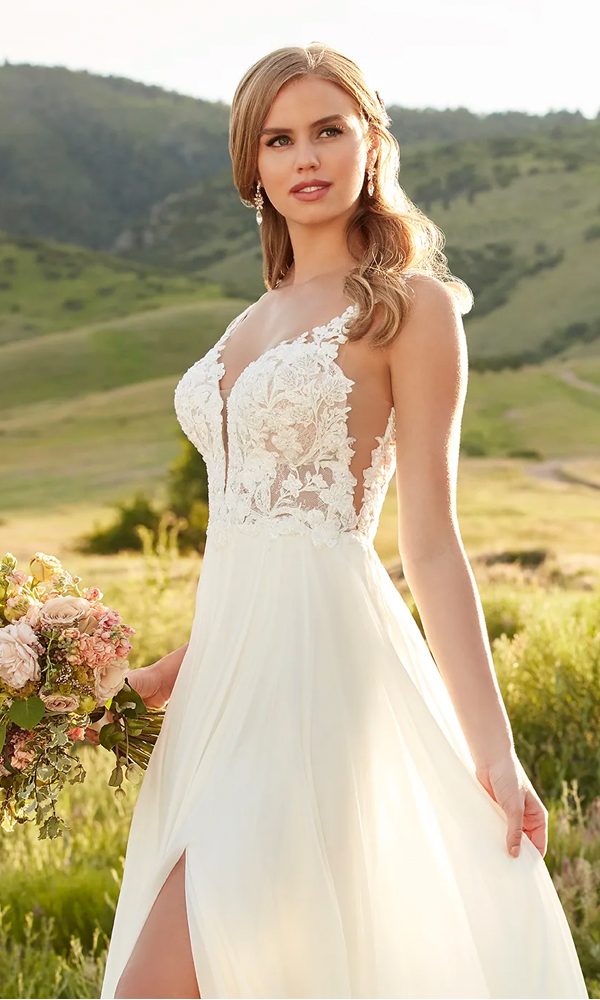 Romantische Braut in einem fließenden Kleid mit tiefem V-Ausschnitt und Spitze, posiert in einer sonnenverwöhnten, hügeligen Landschaft. Der hohe Schlitz im Kleid und die natürliche Umgebung verleihen dem Bild eine leichte, sommerliche Stimmung.