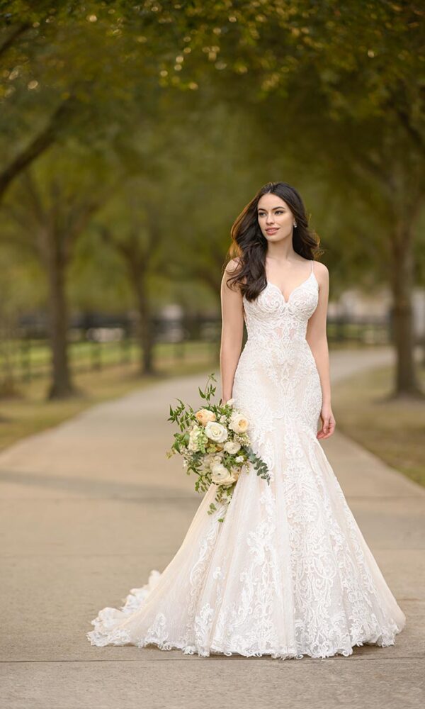 Elegante Braut in einem weißen Spitzenkleid mit floralen Details und zarten Trägern, die mit einem natürlichen Brautstrauß auf einem Weg unter grünen Bäumen posiert.
