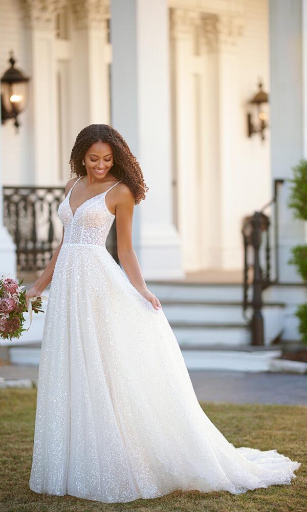 Braut in elegantem, funkelndem weißen Kleid mit zartem Ausschnitt, lächelnd, mit einem Blumenstrauß in der Hand, posiert sie auf einem grünen Weg im Freien.