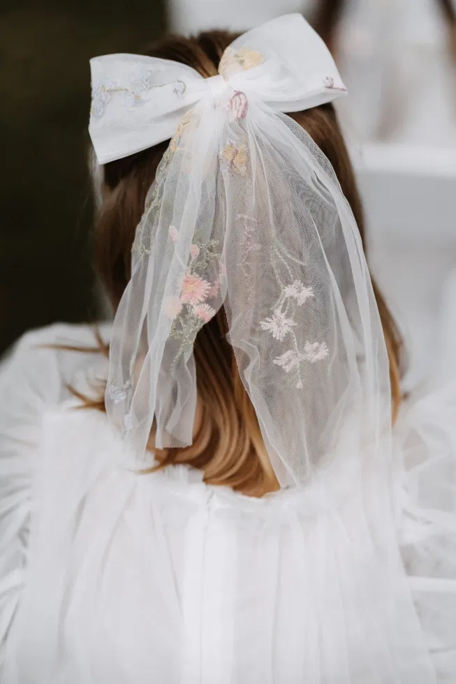 Rückansicht eines Mädchens mit großer weißer Schleife im Haar, an der ein bestickter Tüllschleier mit zarten Blüten hängt.