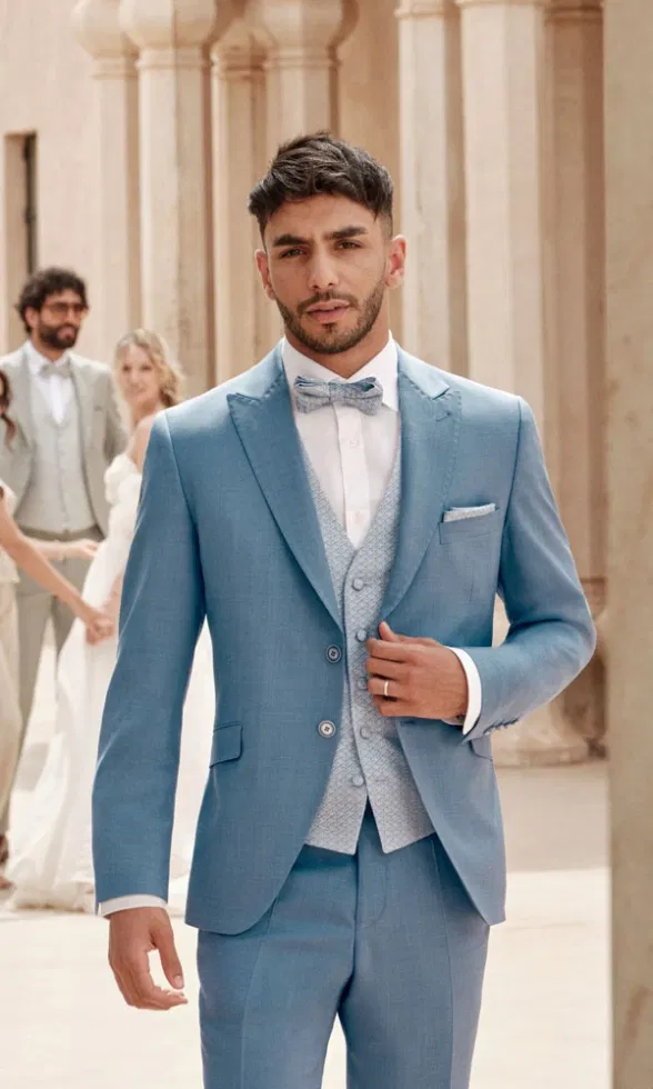 Bräutigam in hellblauem Anzug mit gemusterter Weste und Fliege steht vor sandfarbenen Säulen, während im Hintergrund Hochzeitsgäste und ein Brautpaar zu sehen sind.