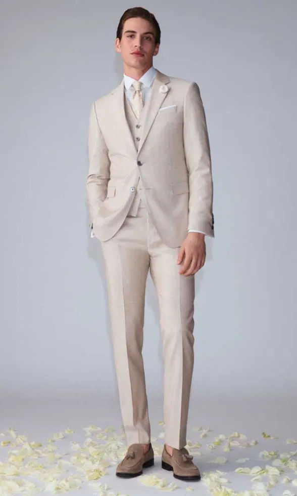 Moderner Bräutigam-Look im Studio: Ein Mann trägt einen hellen Anzug mit Weste, Krawatte und floraler Ansteckblume vor neutralem Hintergrund.