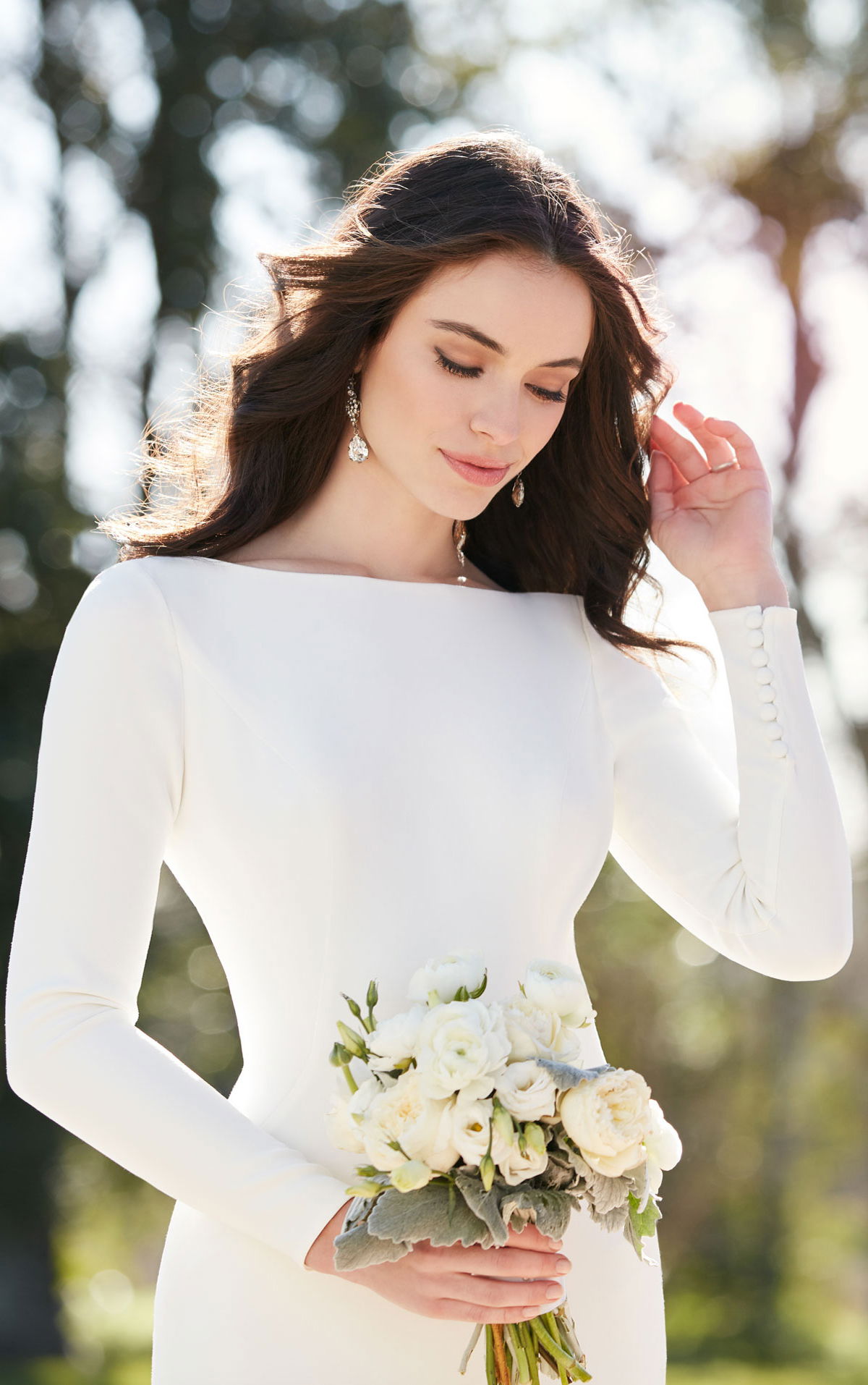 Detailaufnahme einer Braut in einem minimalistischen Hochzeitskleid mit langen Ärmeln und Blumenstrauß in der Hand. Sie schaut nachdenklich nach unten, während das Licht durch die Bäume scheint.