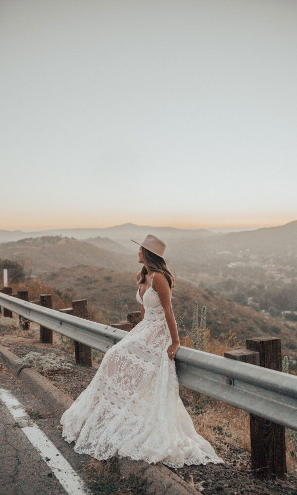 Die Braut in einem Boho-Kleid aus feiner Spitze und Hut sitzt auf einer Leitplanke mit Blick auf eine weite Hügellandschaft im Sonnenuntergang.