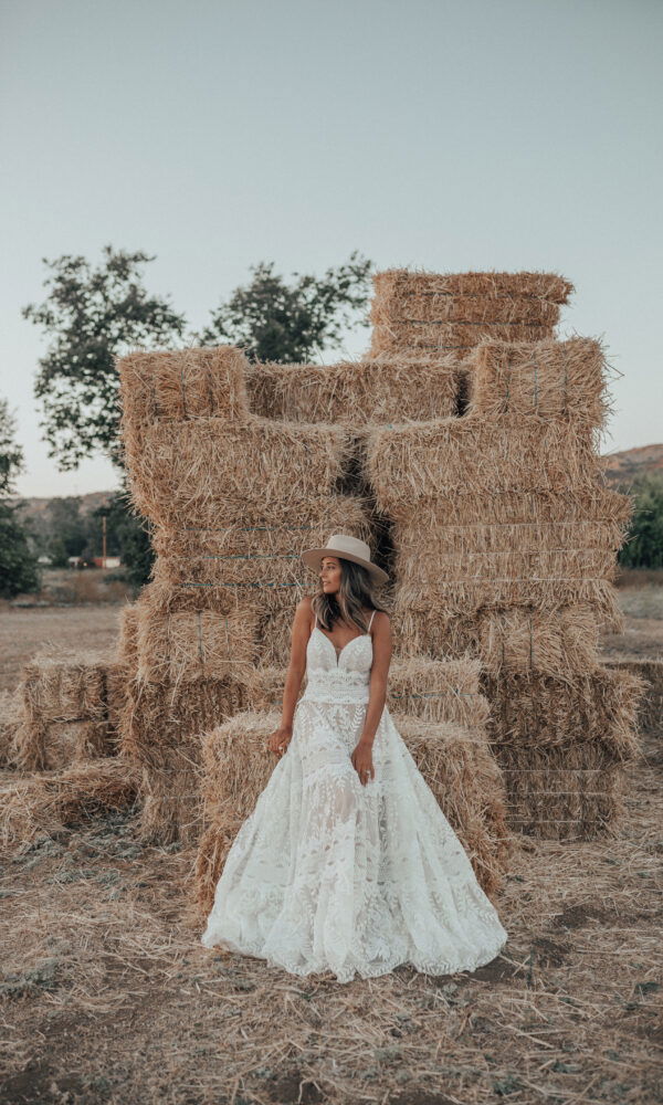 Eine Braut im Boho-Stil mit einem Spitzenkleid und Hut steht vor einer Wand aus Heuballen. Das natürliche Umfeld und das Brautkleid schaffen einen rustikalen, romantischen Look.