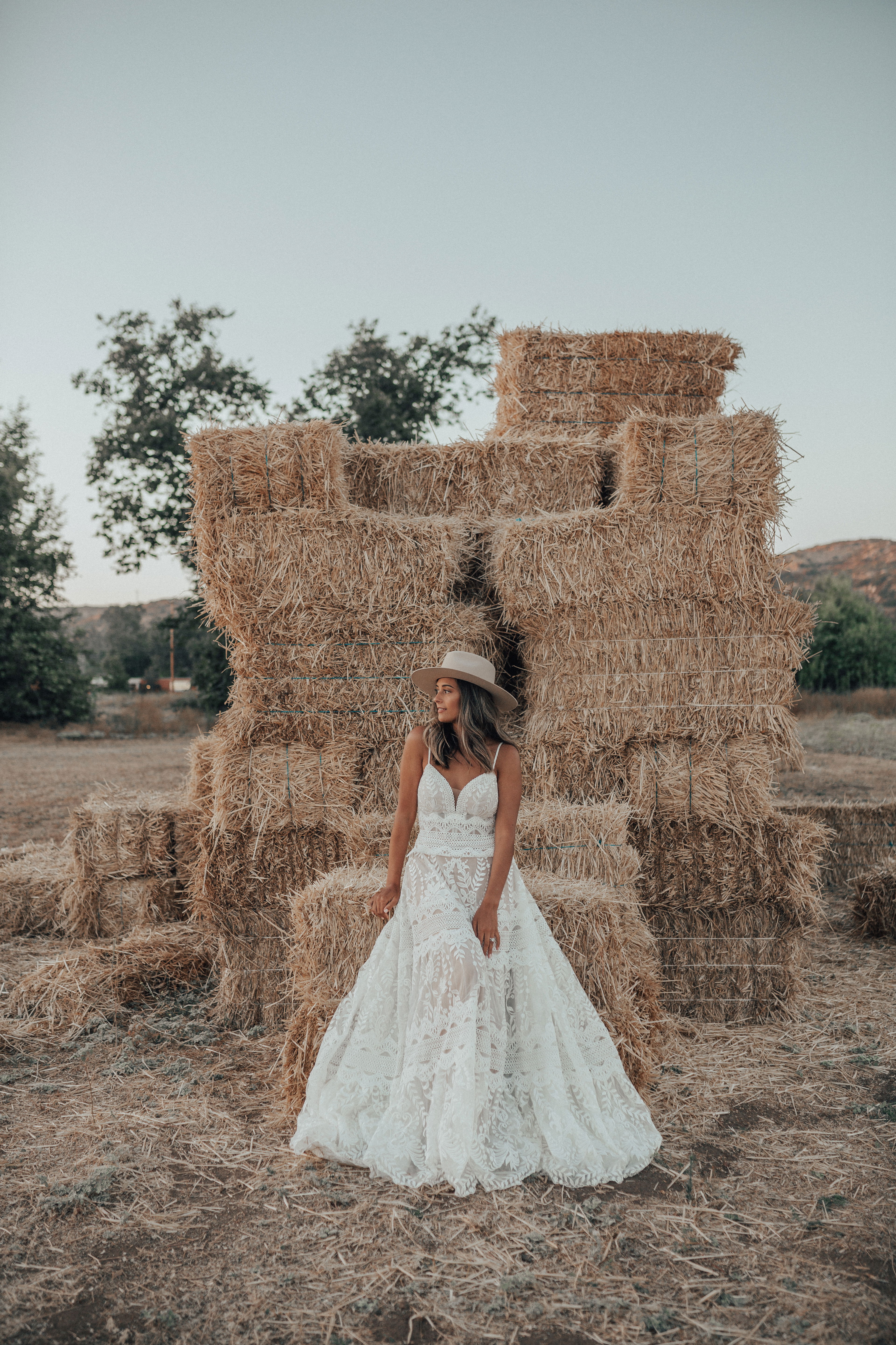 Eine Braut im Boho-Stil mit einem Spitzenkleid und Hut steht vor einer Wand aus Heuballen. Das natürliche Umfeld und das Brautkleid schaffen einen rustikalen, romantischen Look.