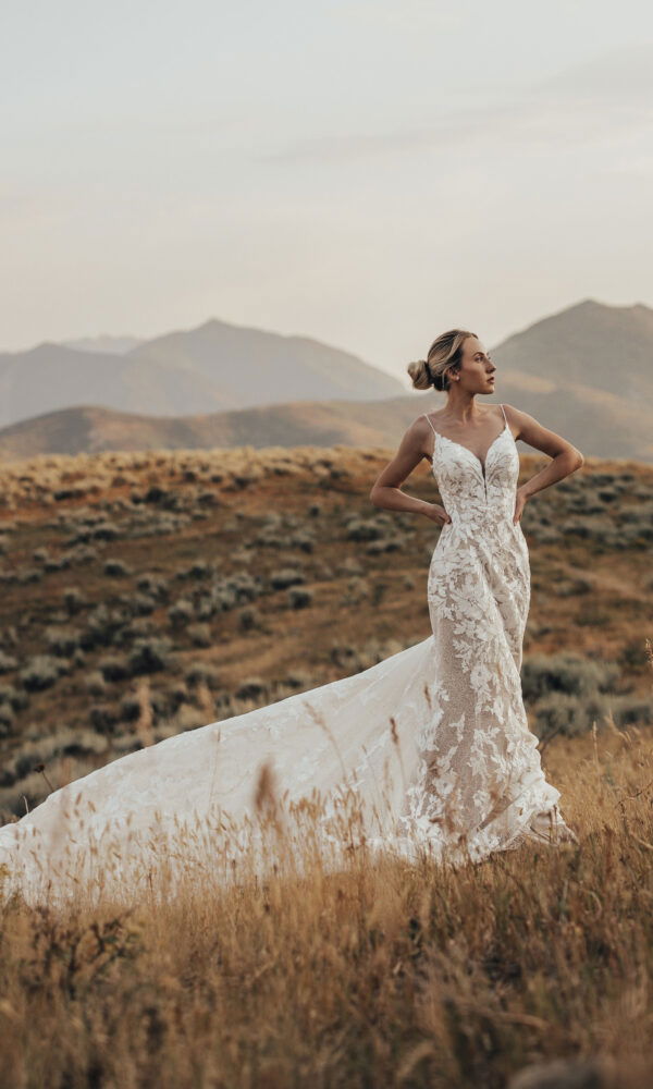 Eine Frau in einem schmal geschnittenen Brautkleid mit floralen Applikationen posiert in einer offenen Feldlandschaft, Berge erheben sich im Hintergrund.