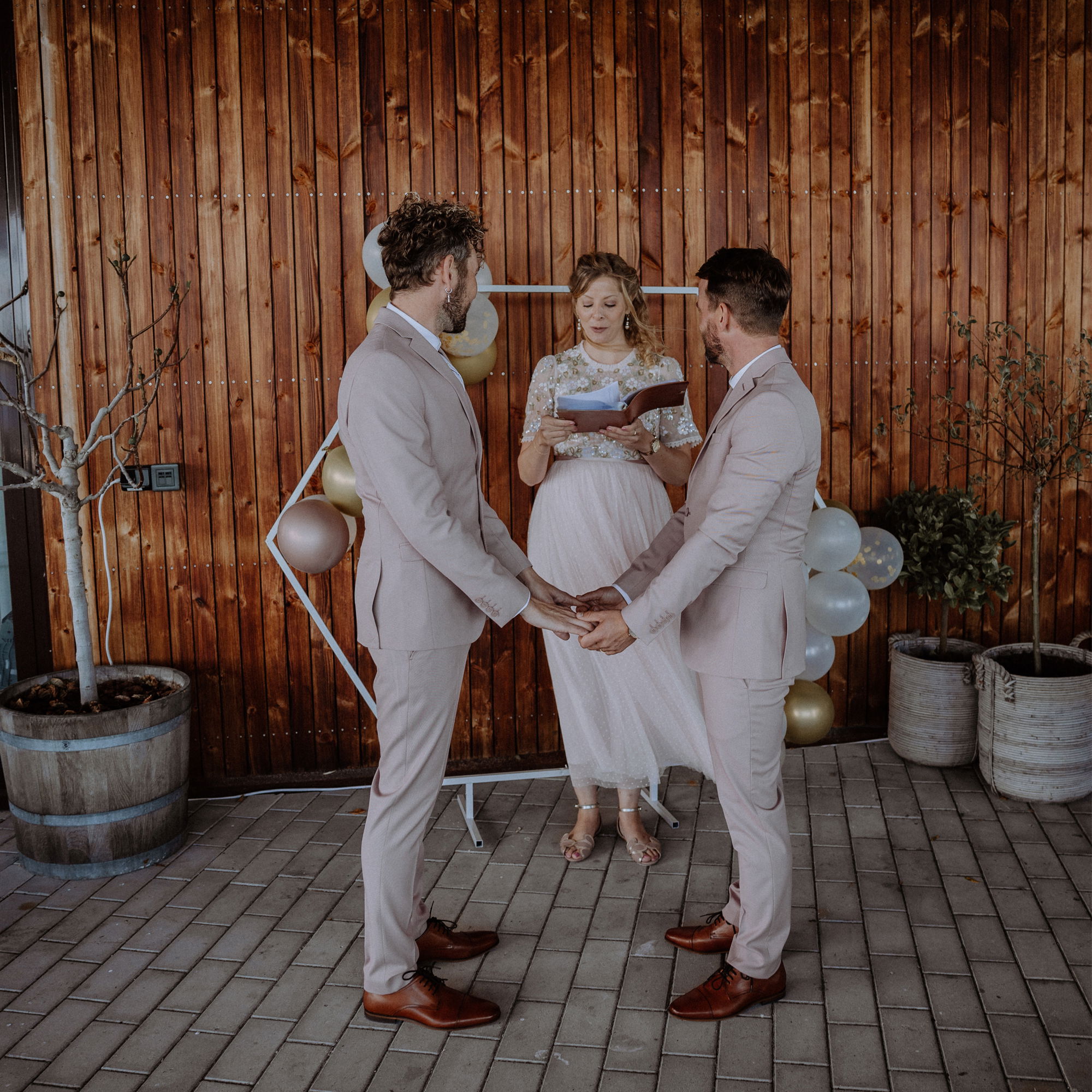 Zwei Männer in rosafarbenen Anzügen stehen Hand in Hand bei ihrer Hochzeit, während die Rednerin das Eheversprechen vorträgt.