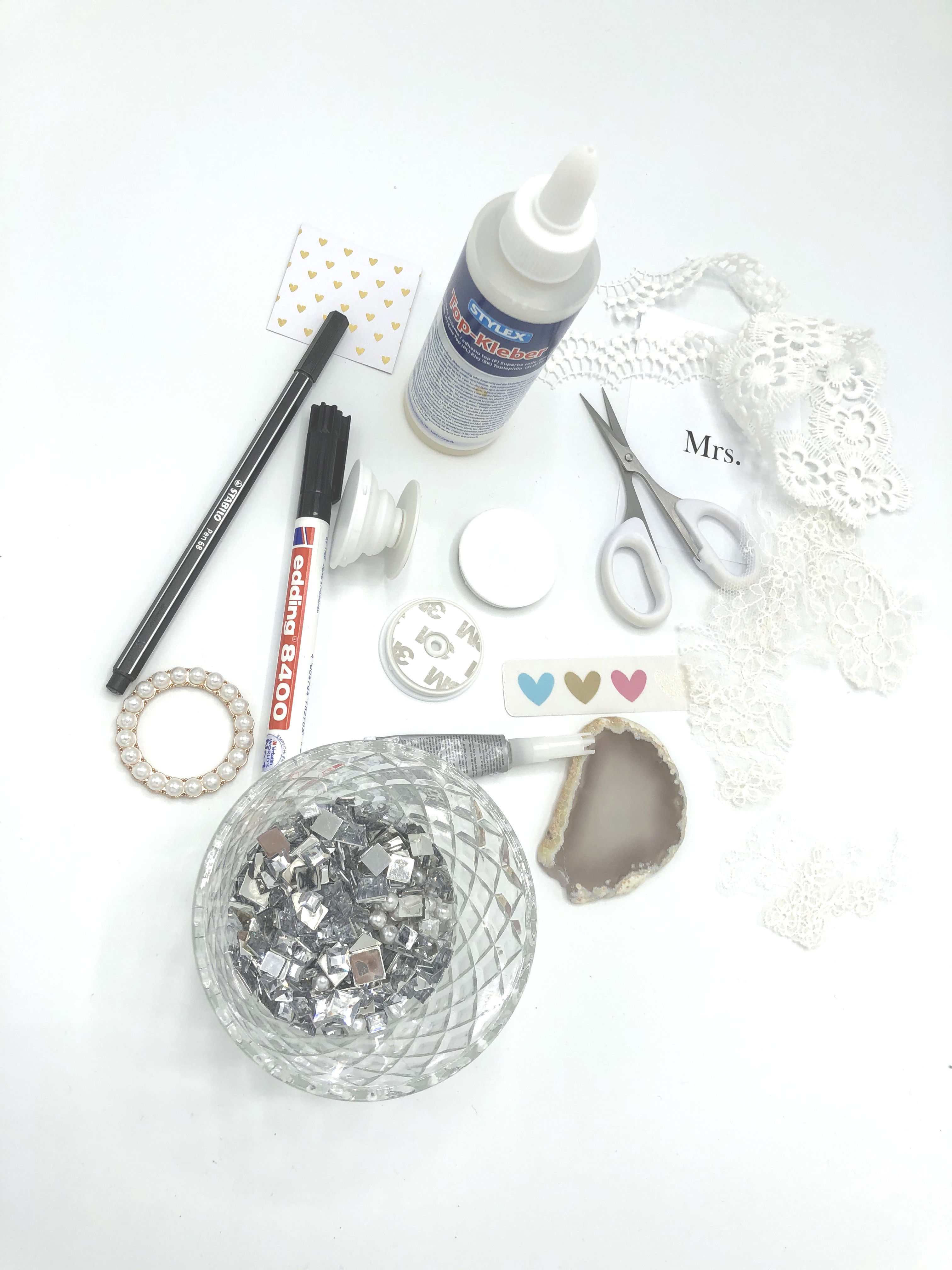 Eine Sammlung von DIY-Bastelutensilien auf einer weißen Unterlage. Spitze, Kleber, eine Schere und bunte Herzaufkleber sind bereit für ein kreatives Projekt.