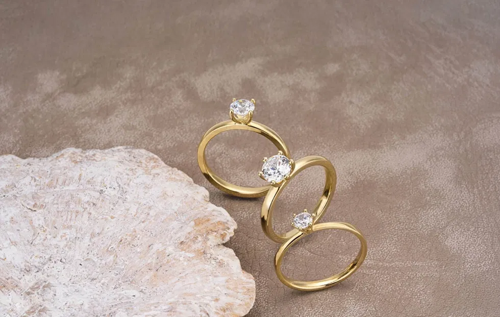 Goldene Solitärringe mit funkelnden Diamanten, arrangiert auf einem hellen Naturstein – minimalistisches Stillleben mit Fokus auf klassische Fassungen.