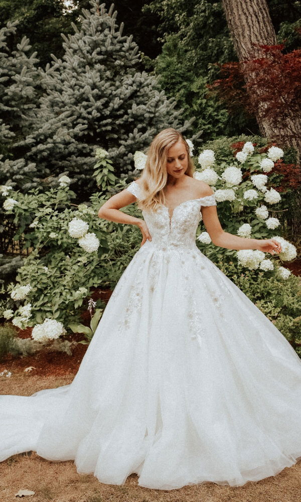 Braut posiert in einem floralen Brautkleid vor üppiger Gartenkulisse. Sie hat eine Hand an der Hüfte und ist umgeben von weißen Hortensien.