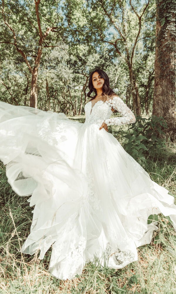 Die Braut in einem Spitzenkleid mit langen Ärmeln posiert im Grünen, die Schleppe des Kleides schwingt im Wind und schafft Bewegung im Bild.