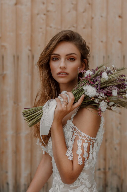 Romantisches Brautporträt im Boho-Stil: Die Braut trägt ein Spitzenkleid und hält einen natürlichen Blumenstrauß, während sie verträumt in die Kamera blickt.