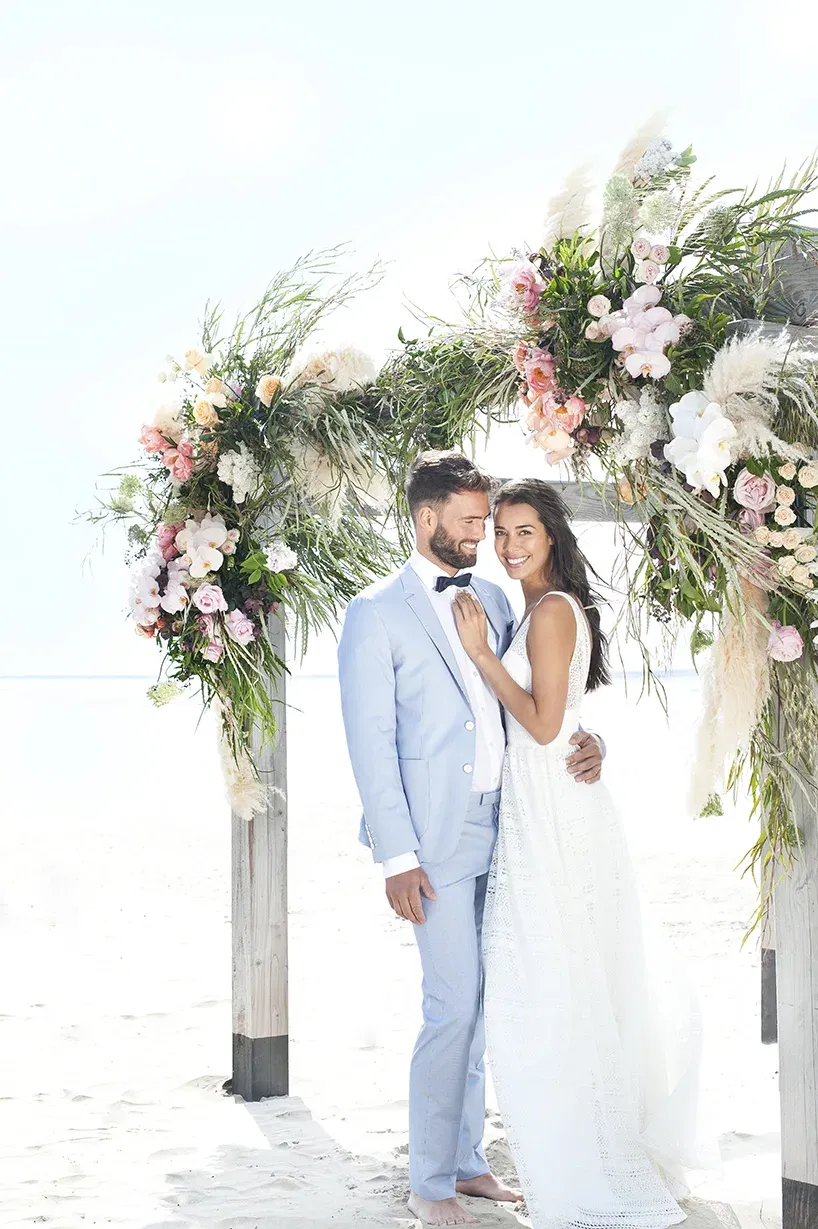 Romantisches Strandfoto eines frisch verheirateten Paares, das sich unter einem blumigen Traubogen in natürlichen Farben befindet.
