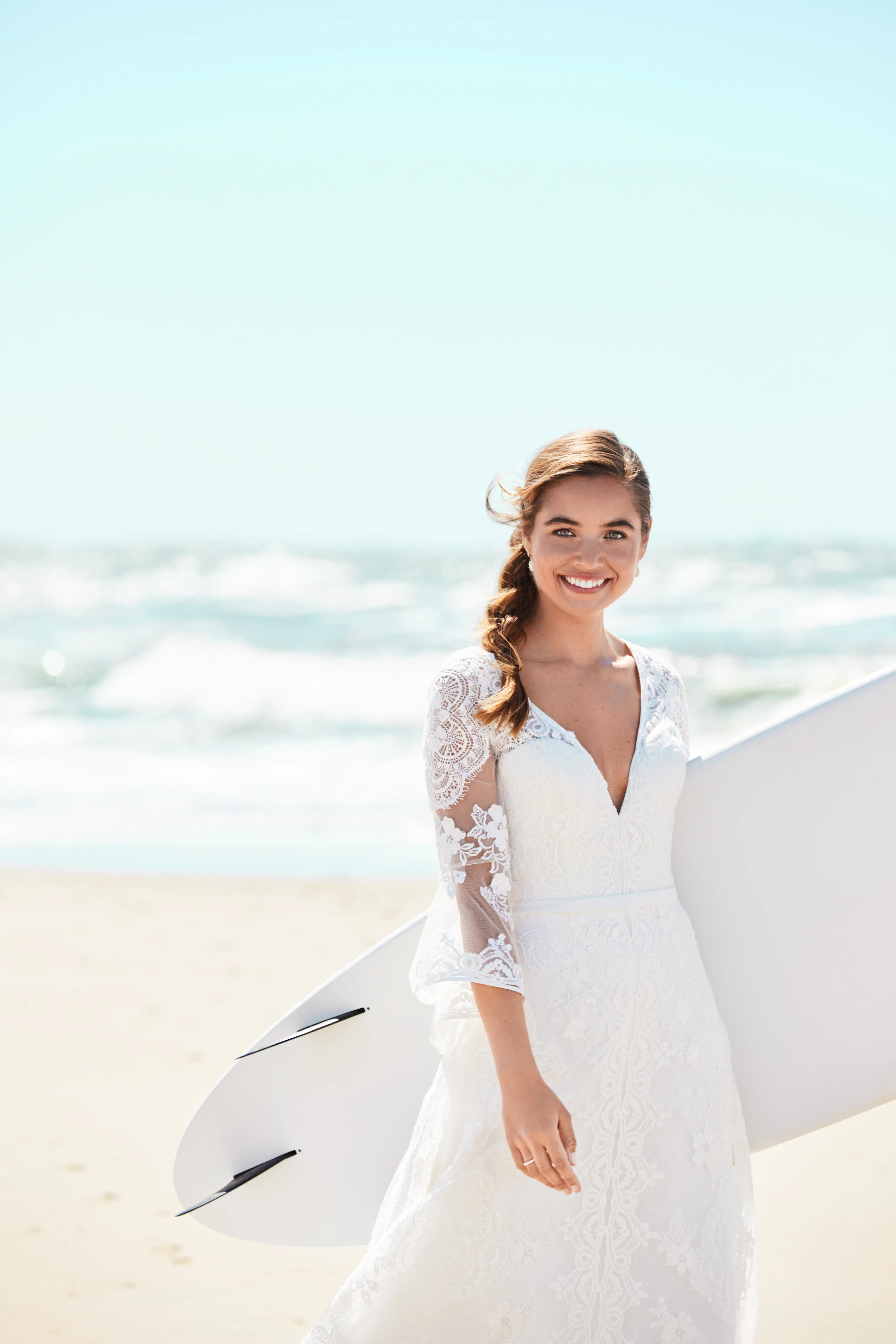 Braut mit Surfbrett und Spitzenkleid am Strand, lächelnd in die Kamera, während die Wellen im Hintergrund sanft ans Ufer rollen.