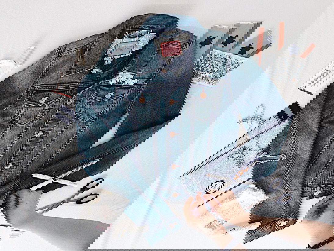 Jeansjacke im Flatlay-Stil mit kreativen Bastelmaterialien wie Glitzerfolie, Perlen und einer Schere. Zwei Hände beginnen mit der Verzierung der Jacke, um sie individuell zu gestalten.