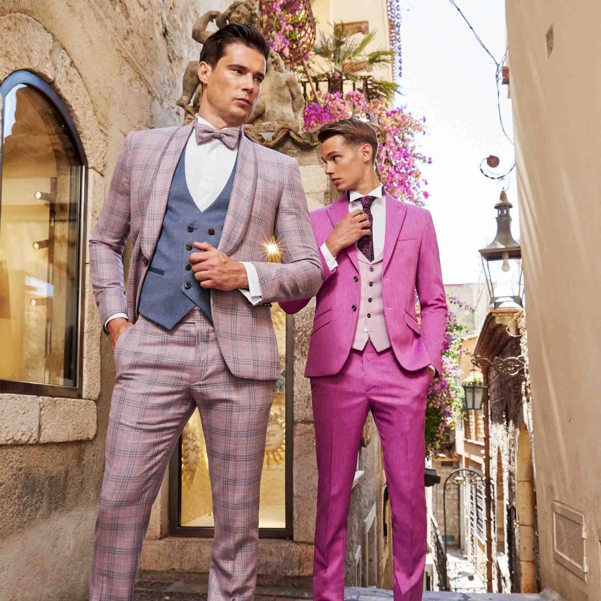 Zwei Männer in eleganten, farbigen Anzügen – einer in kariertem Rosé-Grau, der andere in kräftigem Pink – posieren stilvoll in einer mediterranen Gasse mit Blüten und historischen Fassaden.