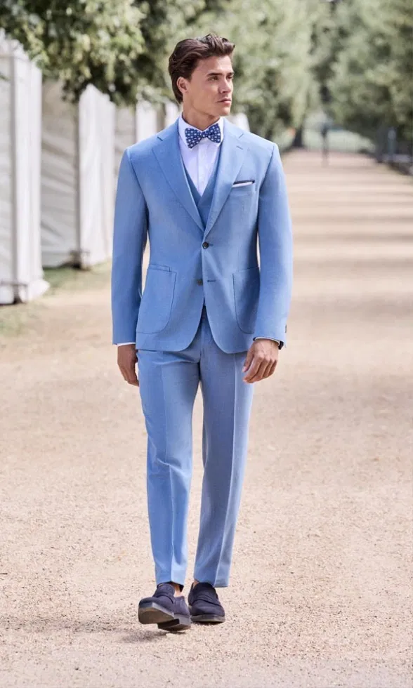 Mann in hellblauem Anzug mit passender Weste und gepunkteter Fliege geht einen Kiesweg entlang, umgeben von Bäumen und Zelt