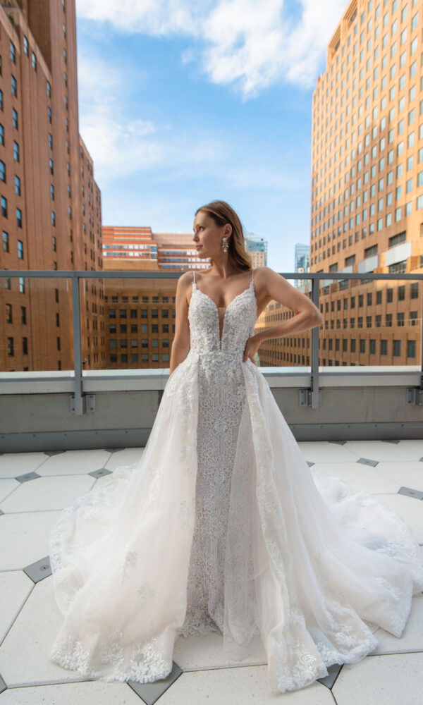 Die Braut posiert auf einer Dachterrasse mit spektakulärer Aussicht auf die Skyline. Ihr elegantes Spitzenkleid mit tiefem Ausschnitt und langen Schleppe strahlt in der Sonne.