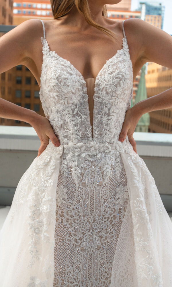 Nahaufnahme des aufwendig verzierten Brautkleides mit floraler Spitze und tieferem Dekolleté. Die Braut betont die Details durch ihre Hände an der Taille, was die hochwertige Verarbeitung hervorhebt.