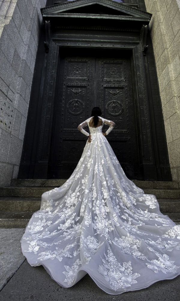 Rückansicht einer Braut in einem eleganten Kleid mit Blumenstickerei und langer Schleppe, die vor einer massiven schwarzen Tür posiert und die Hände in die Hüften stützt.