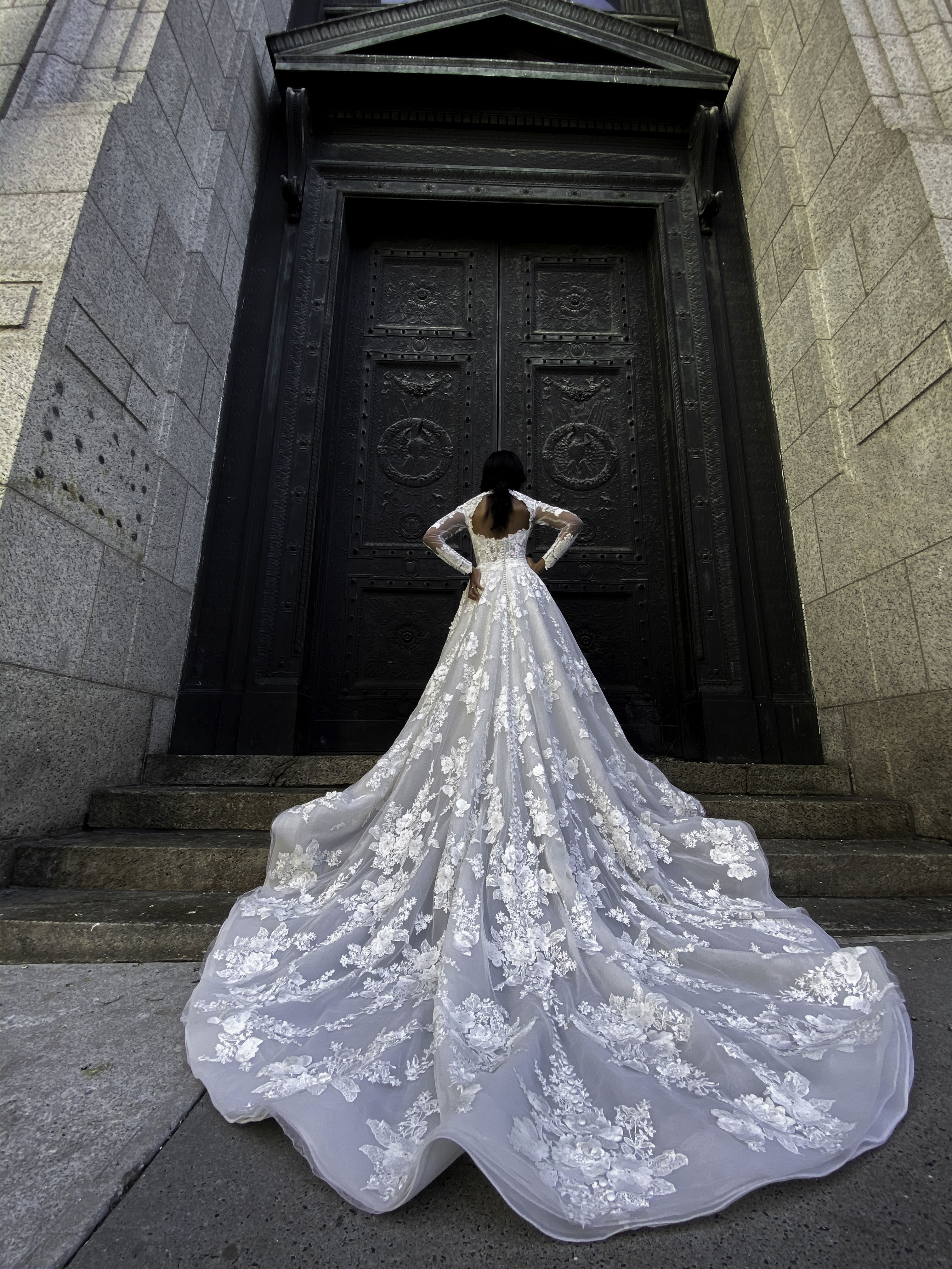 Rückansicht einer Braut in einem eleganten Kleid mit Blumenstickerei und langer Schleppe, die vor einer massiven schwarzen Tür posiert und die Hände in die Hüften stützt.