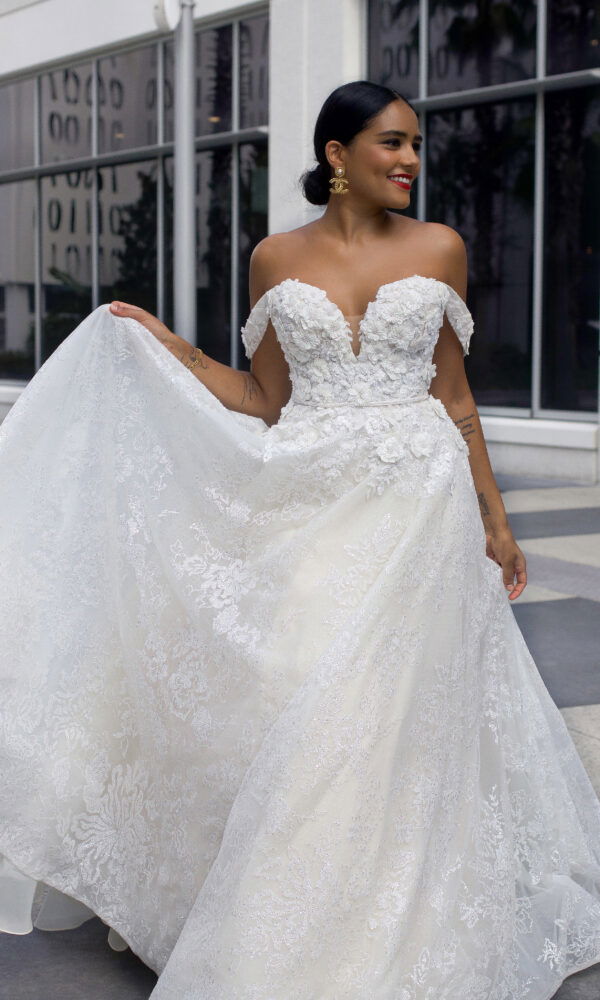 Elegante Braut in einem trägerlosen Kleid mit aufwendiger Spitzenverzierung und Blumenelementen, lächelt vor urbaner Kulisse.