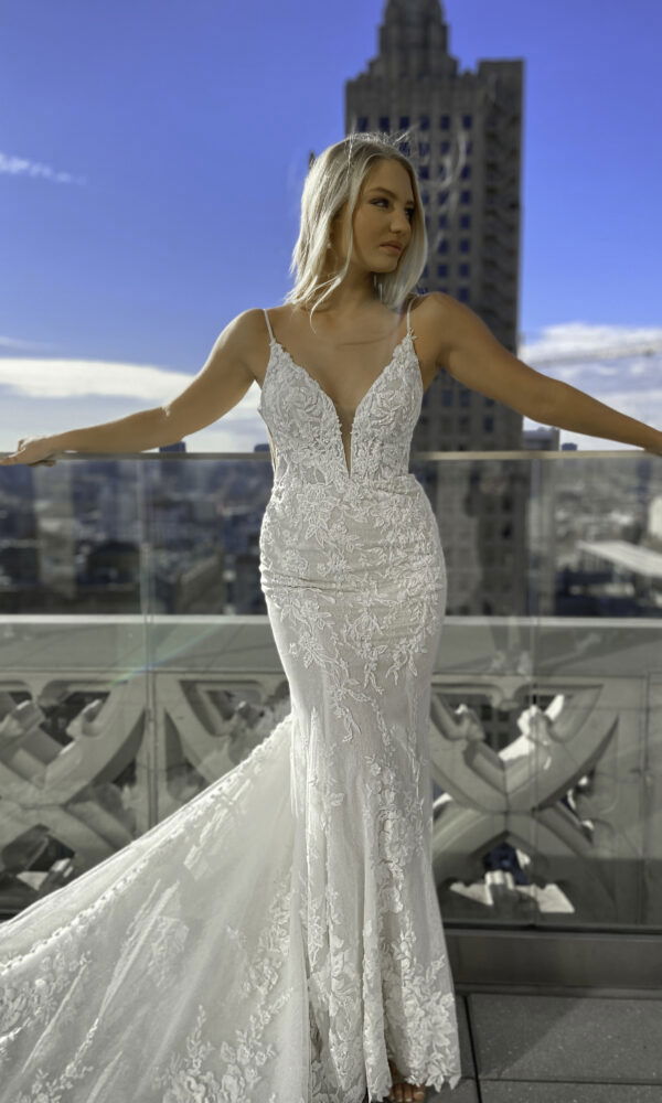 Die Braut in einem eleganten, spitzenbesetzten Kleid posiert auf einem Stadtbalkon, Arme auf dem Geländer ruhend, während der klare Himmel und die Skyline das Bild ergänzen.