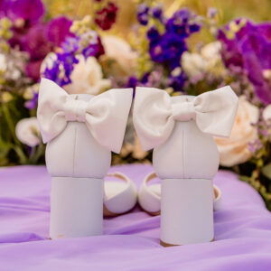 Weiße Hochzeitsschuhe mit massiven Absätzen und auffälligen Schleifen auf der Rückseite stehen auf einem lila Stoff. Dahinter ragen bunte Blumen, hauptsächlich in Lila- und Rosatönen, hervor, was dem Bild einen romantischen und eleganten Touch verleiht, passend für eine stilvolle Hochzeit.