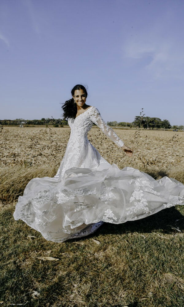 Lächelnde Braut in einem eleganten Spitzenkleid dreht sich auf einem offenen Feld, während der Rock des Kleides sich dynamisch bewegt.