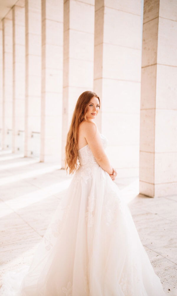 Eine junge Frau im Brautkleid posiert in einem lichtdurchfluteten Raum mit hohen Säulen. Sie hat langes, welliges Haar und schaut sanft zur Seite.