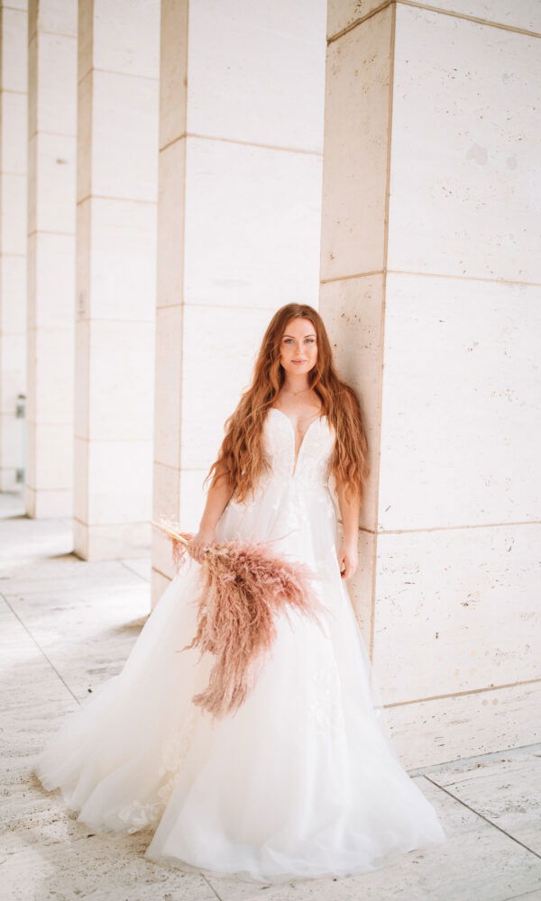 Junge Frau in einem eleganten Brautkleid mit tiefem Dekolleté posiert vor einer hellen Marmorwand. Sie hat lange, rote Haare und hält einen Strauß aus Pampasgras. Das weiche Licht und die schlichte Architektur unterstreichen die ruhige Szene.