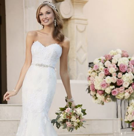 Elegante Braut in einem figurbetonten, spitzenbesetzten Kleid von Stella York, geschmückt mit einer Tiara, posiert vor prachtvollen Blumenbouquets.