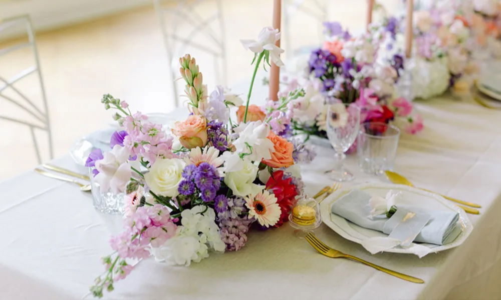 Hochzeitstisch mit eleganter Blumendekoration in Pastelltönen, goldfarbenem Besteck, weißen Tellern und zart gefaltetem Servietten-Arrangement.