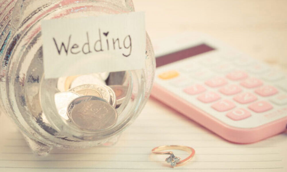 Sparglas mit der Aufschrift 'Wedding' neben einem rosa Taschenrechner und einem goldenen Verlobungsring – Finanzplanung für die Hochzeit.