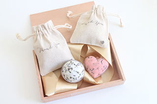 Stilvolle Geschenkverpackung mit zwei naturfarbenen Stoffbeuteln, bedruckt mit "Thank You", und zwei handgemachten Badekugeln – eine rund mit Blüten, die andere herzförmig.