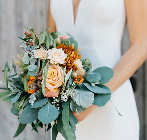 Eine Braut in einem eleganten weißen Kleid hält einen üppigen Blumenstrauß mit Pfirsichrosen, gelben Chrysanthemen und grünen Eukalyptusblättern.