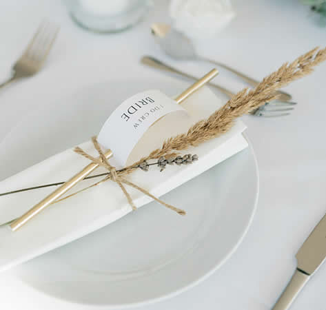 Eine minimalistische Tischdekoration mit einem weißen Teller, einer Serviette, einer Namenskarte mit der Aufschrift 'Bride' und einer kleinen Trockenblume, umwickelt mit Juteband.