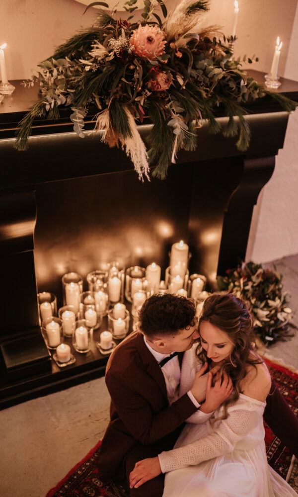 Das Brautpaar sitzt vor einem Kamin, umgeben von Dutzenden Kerzen und Blumenarrangements, die eine romantische, weihnachtliche Stimmung erzeugen.