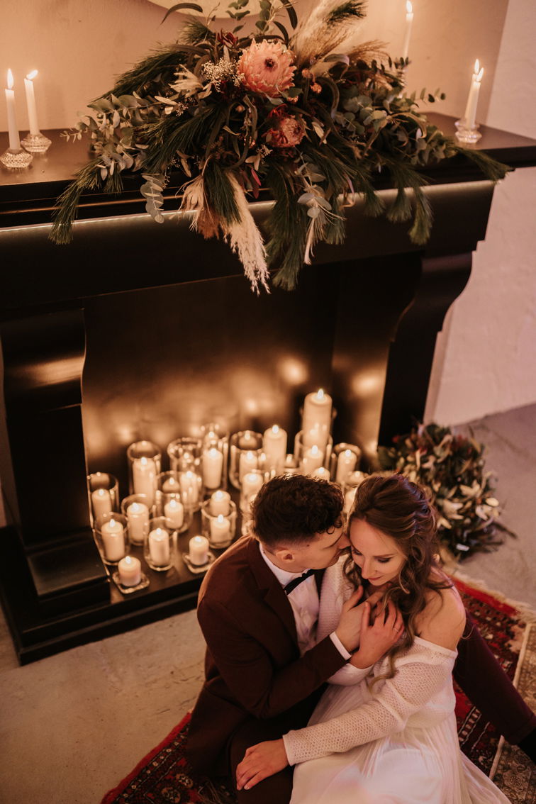 Das Brautpaar sitzt vor einem Kamin, umgeben von Dutzenden Kerzen und Blumenarrangements, die eine romantische, weihnachtliche Stimmung erzeugen.