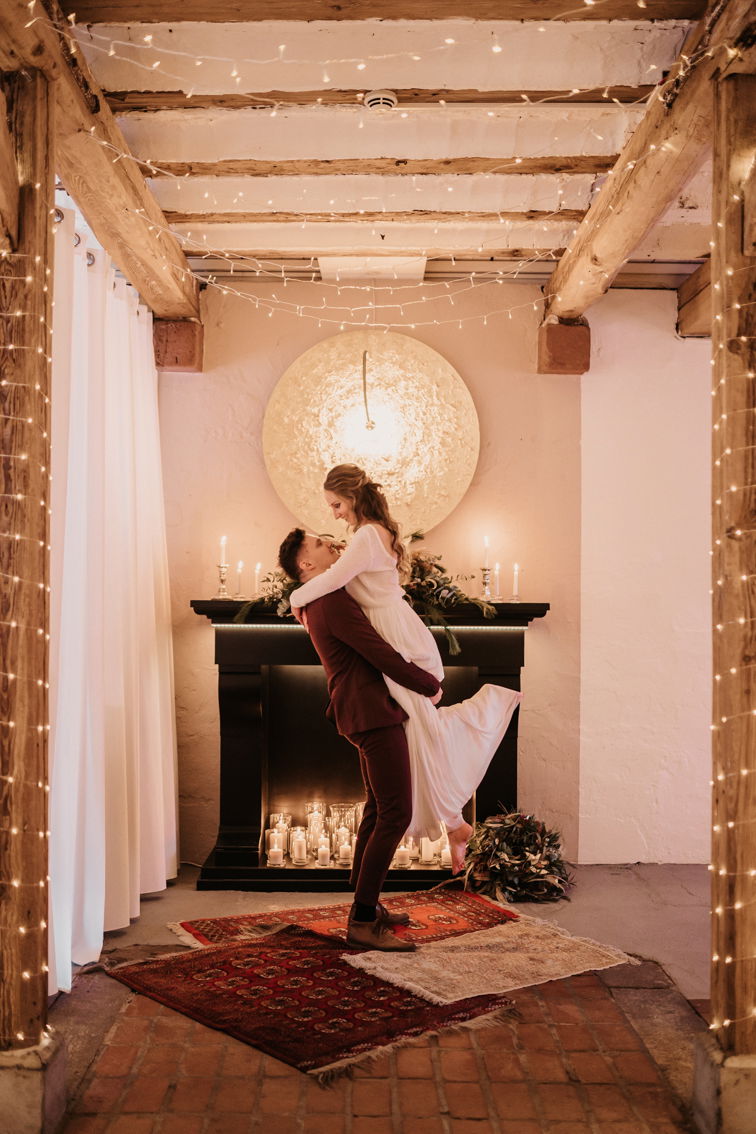 Bräutigam hebt seine Braut vor einem festlich geschmückten Kamin in die Höhe, umgeben von warmen Lichtern und einem gemütlichen Ambiente.