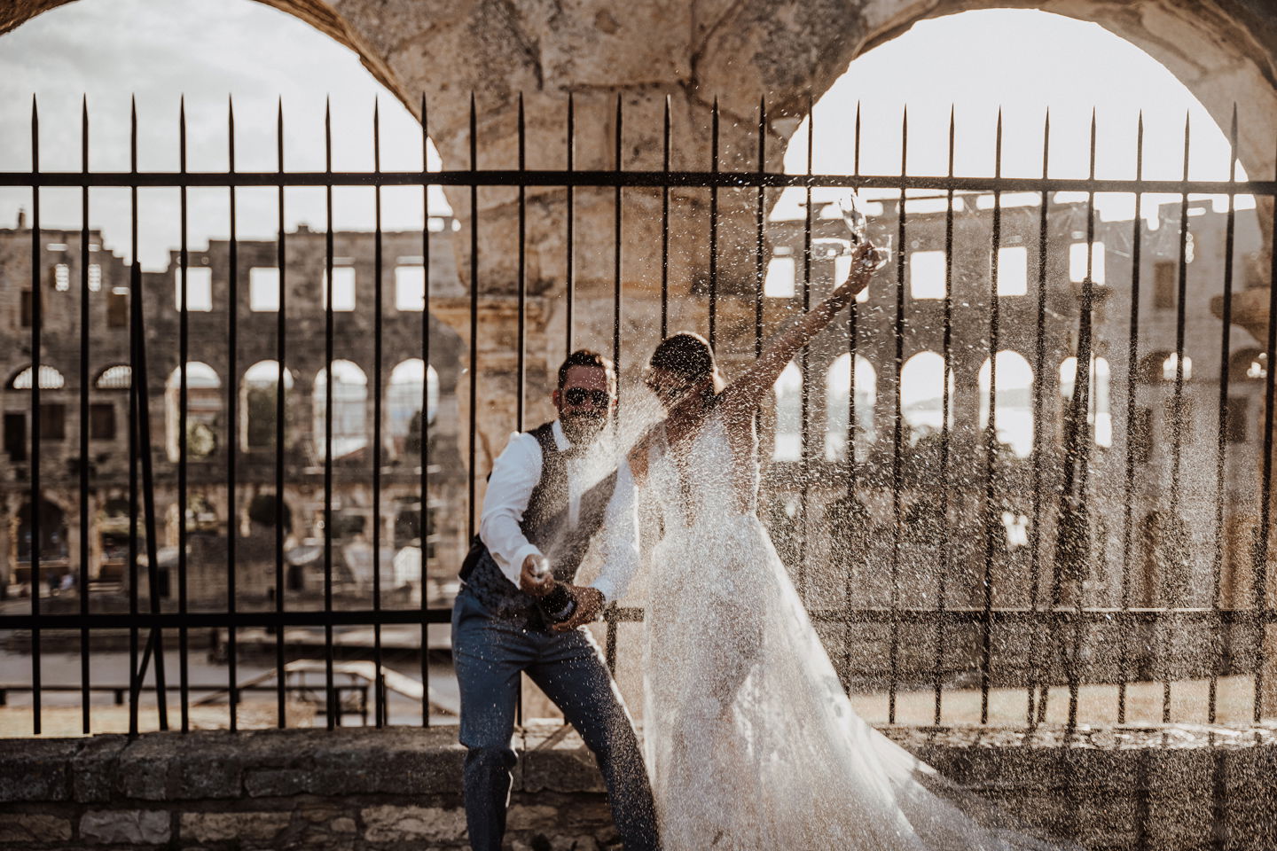 Das Brautpaar sprüht Champagner in die Luft, während sie jubeln. Die Braut in einem weißen Kleid und der Bräutigam in Weste und Sonnenbrille feiern ausgelassen mit Champagner.