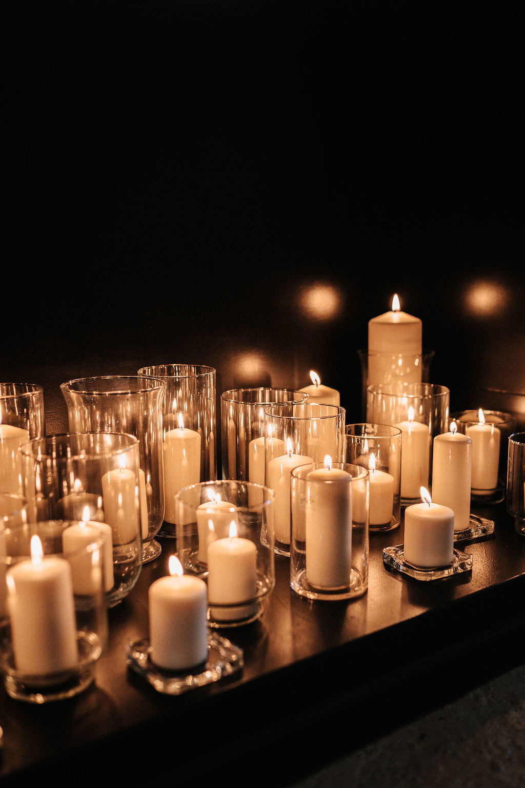 Ein Arrangement aus verschiedenen weißen Kerzen in Gläsern, die in einem dunklen Raum für eine romantische und festliche Stimmung sorgen.