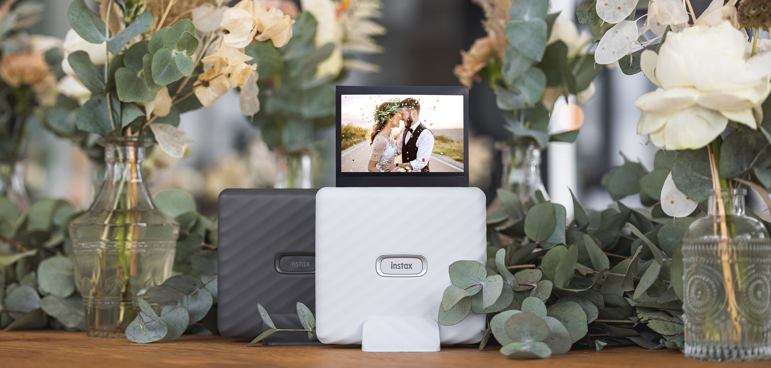 Weisser und schwarzer Instax Link Wide Printer auf einem Tisch, dekoriert mit Eukalyptuszweigen und Trockenblumen. Ein romantisches Hochzeitsfoto eines sich küssenden Paares ist auf dem weissen Drucker zu sehen. Die dezenten Blumen und Blätter schaffen eine natürliche und elegante Szenerie.