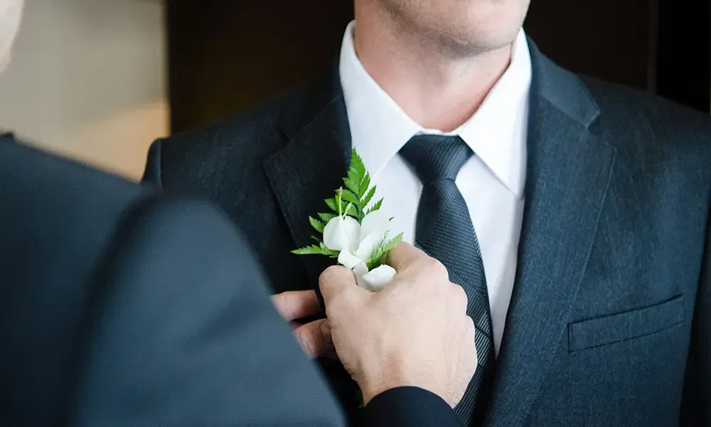 Ein Mann im Anzug wird für die Hochzeit vorbereitet, indem ihm eine weiße Boutonniere mit grünen Blättern angesteckt wird.