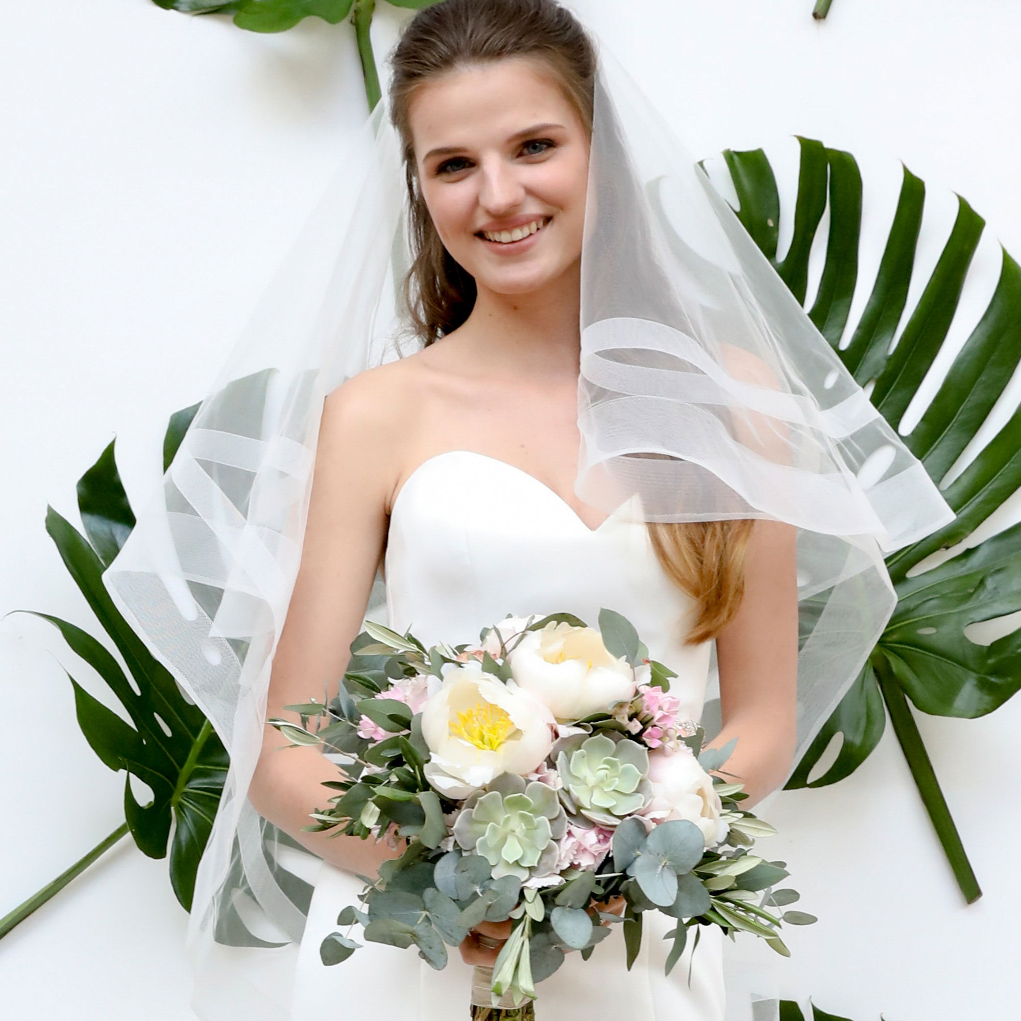 Junge Braut mit einem weißen, trägerlosen Kleid und einem üppigen Blumenstrauß, bestehend aus Sukkulenten und weißen Blumen. Sie steht vor einem Hintergrund mit großen Monstera-Blättern und trägt einen zarten, durchsichtigen Schleier.