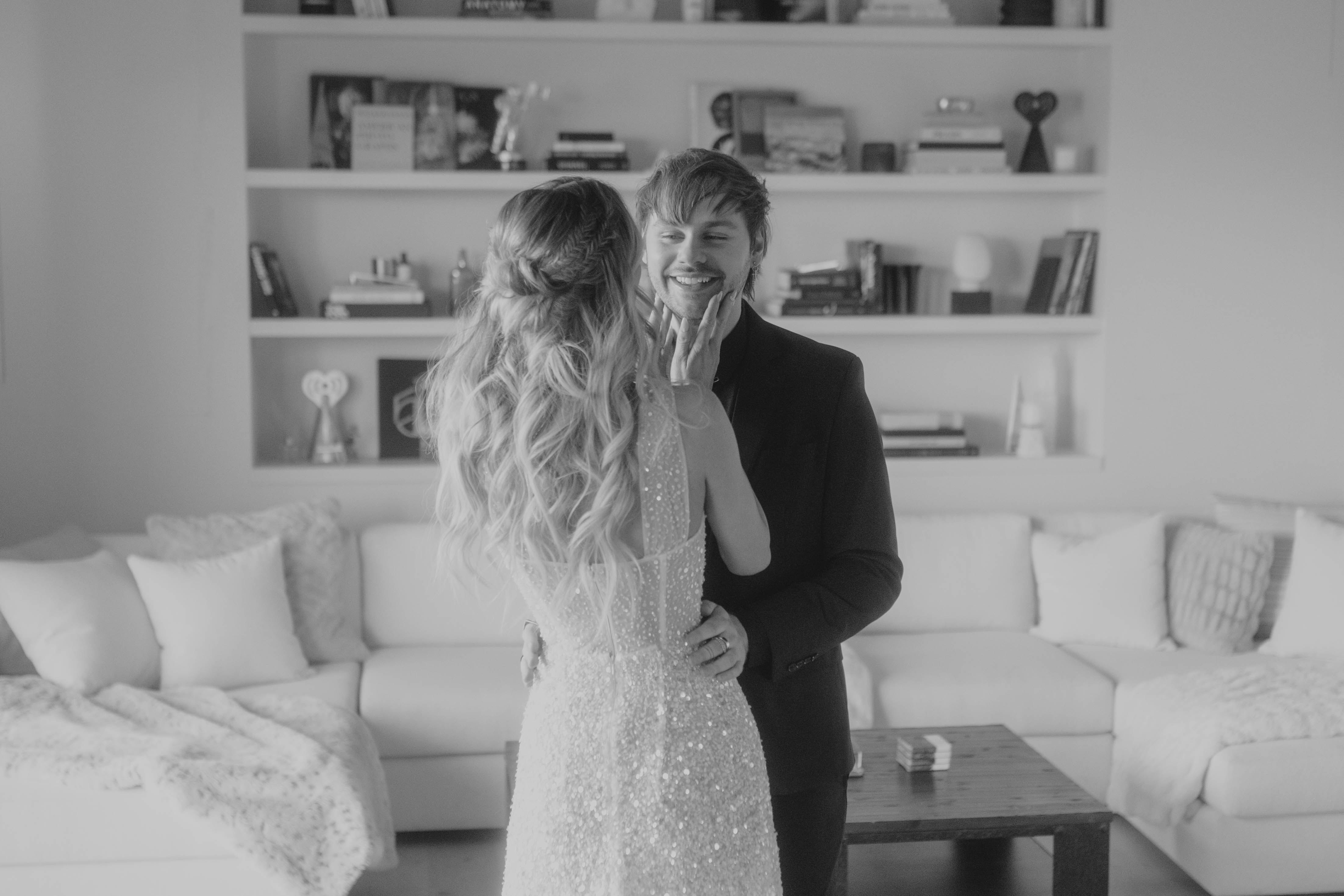 Eine Frau in einem glitzernden Kleid steht vor einem Mann in einem schwarzen Anzug und berührt liebevoll sein Gesicht. Sie befinden sich in einem modernen Wohnzimmer.