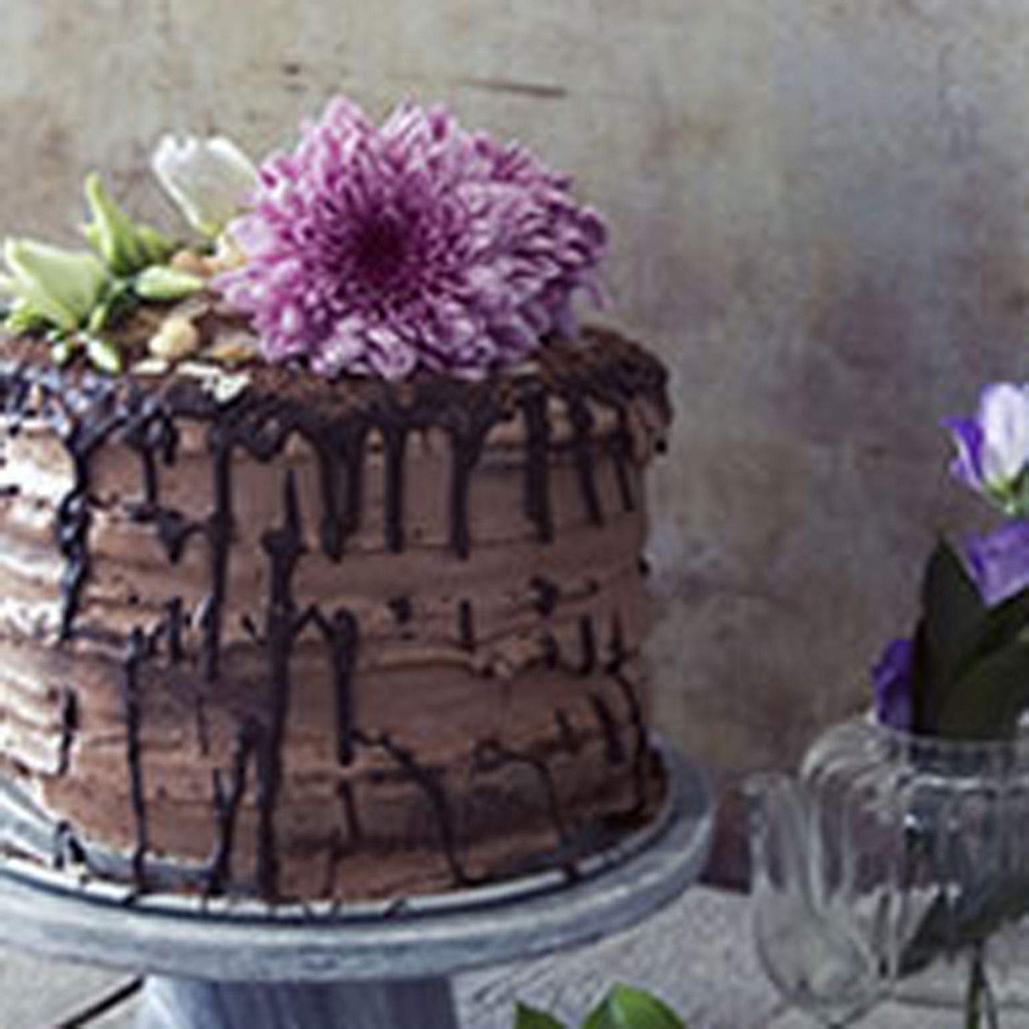Schichttorte mit Schokoganache und fließender Glasur, verziert mit einer großen Blüte, umgeben von zarten lila Blumen in eleganten Vasen.