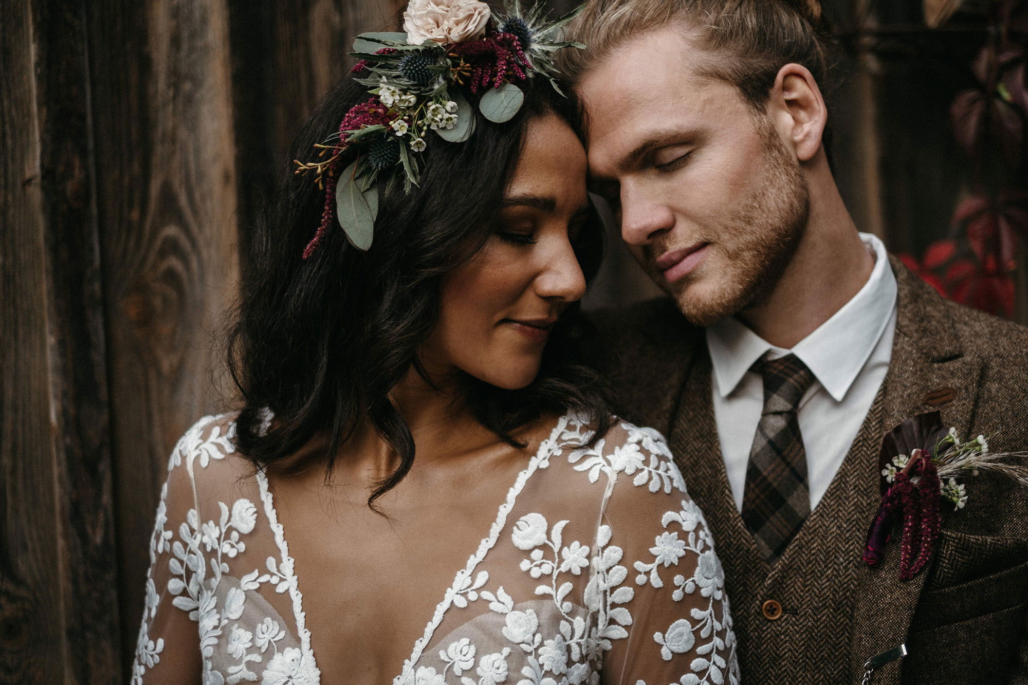 Ein Brautpaar lehnt sich zärtlich aneinander, die Braut trägt ein Spitzenkleid und einen Blumenkranz mit Eukalyptus und Beeren, der Bräutigam einen braunen Tweedanzug.